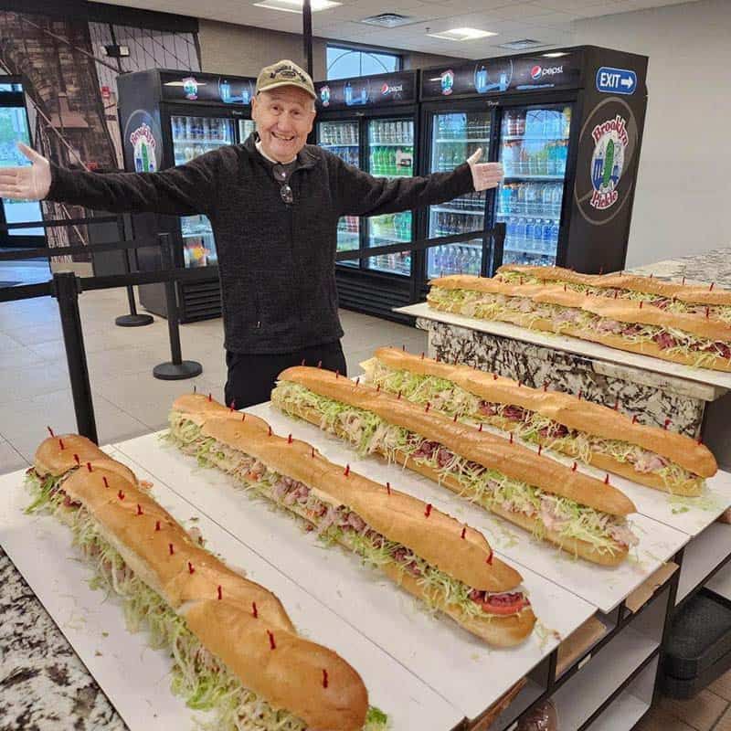 Brooklyn Pickle team member with four three-foot party subs on the counter